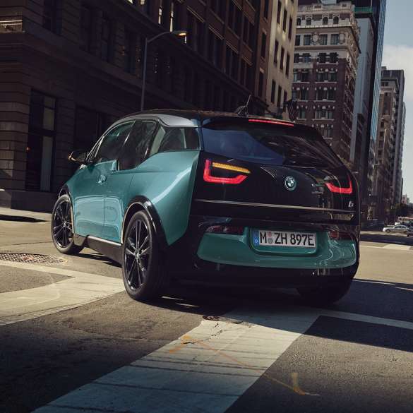 BMW i3 2018 sedd snett bakifrån körandes BMW i3 I01 2018 Jucarobeige med accent Frozen Grey metallic sedd snett bakifrån körandes i stadskuliss
