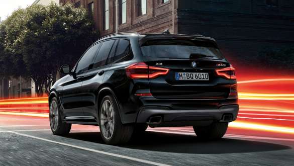 BMW X3 M40i, Trefjärdedels vy bakifrån. BMW X3 M40i i Safirsvart metallic, kommer körande, Exteriör, trefjärdedels vy bakifrån framför stadskuliss med ljusstrimmor.