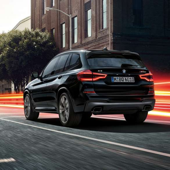 BMW X3 M40i, Trefjärdedels vy bakifrån. BMW X3 M40i i Safirsvart metallic, kommer körande, Exteriör, trefjärdedels vy bakifrån framför stadskuliss med ljusstrimmor.