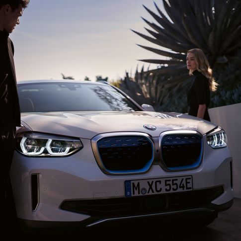 BMW iX3 G08 sedd snett framifrån med BMW i njurar BMW iX3 G08 2020 Mineralvit metallic sedd snett bakifrån med kvinna med BMW i njurar