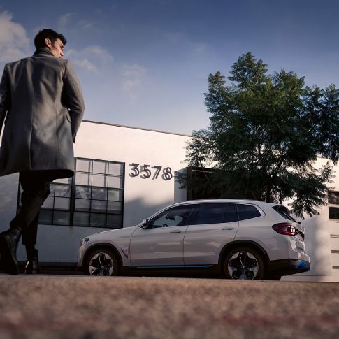 BMW iX3 G08 sedd från sidan framför byggnad med man BMW iX3 G08 2020 Mineralvit metallic vy från sidan framför byggnad med man