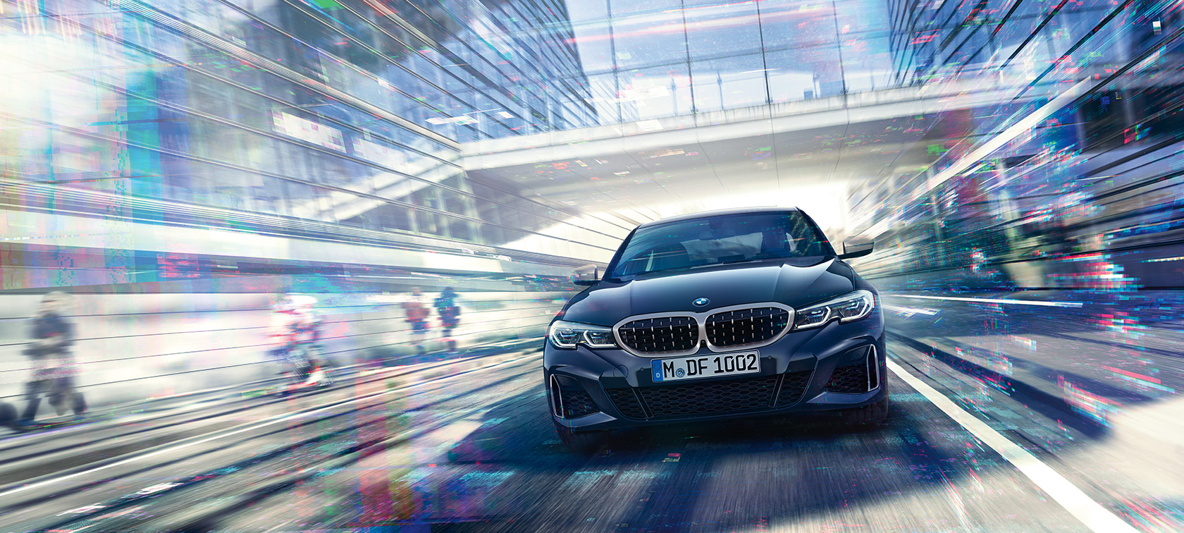 BMW 3-serie Sedan, BMW M340i xDrive framifrån. BMW M340i xDrive Sedan i rörelse sedd framifrån mellan glasfasader i en storstad.