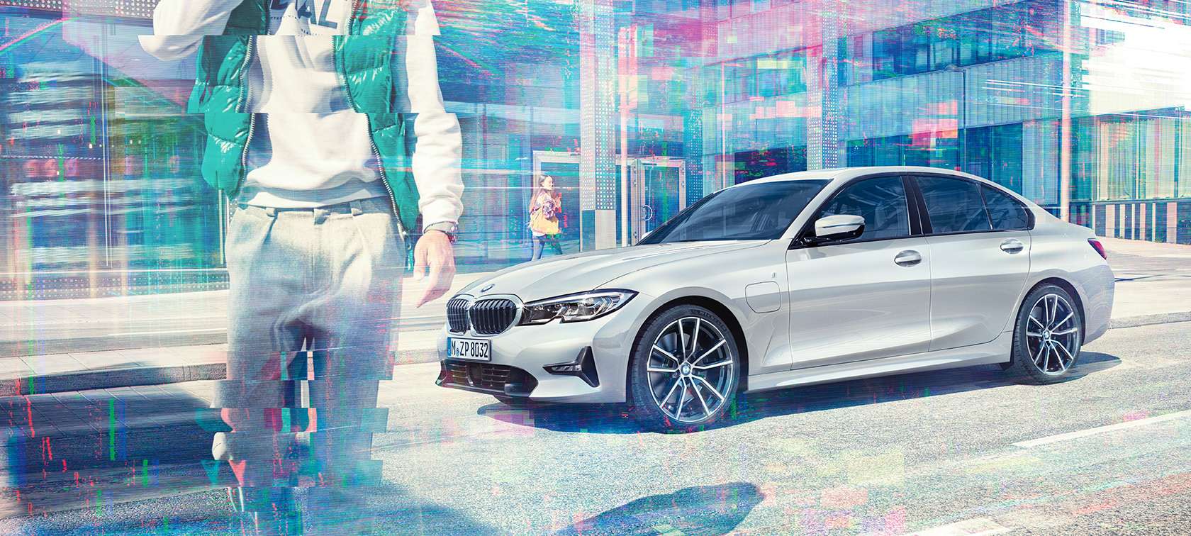 BMW 330e Sedan, från sidan. Stillastående BMW 330e Sedan snett från sidan framför urban glasfasad med förbipasserande människor.