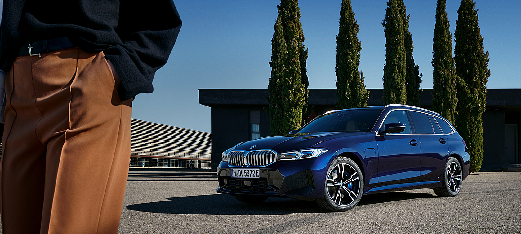 BMW 3-serie Touring LCI G21 Tansanitblå II sedd snett framifrån parkerad framför träd