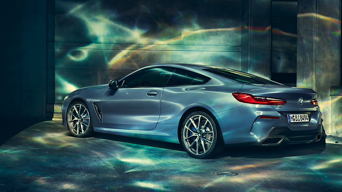 BMW 8-serie Coupé, trefjärdedelsvy bakifrån. Trefjärdedelsvy bakifrån av BMW 8-serie Coupé på natten framför stadsmiljö.