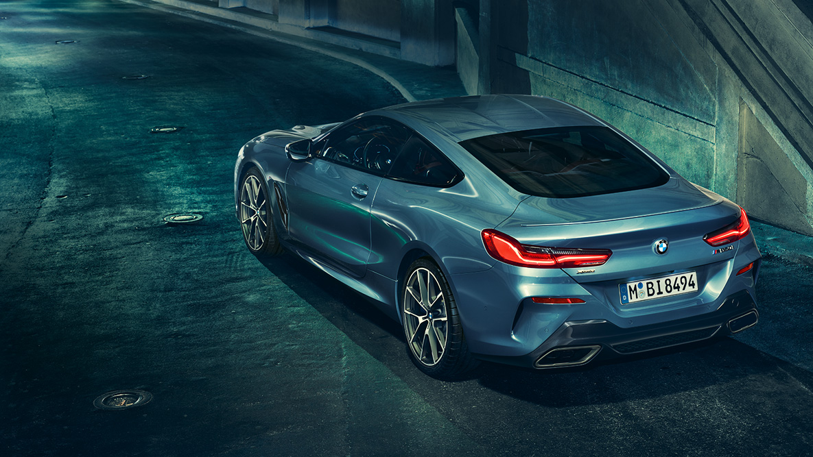 BMW 8-serie Coupé, trefjärdedelsvy bakifrån. Trefjärdedelsvy bakifrån av BMW 8-serie Coupé på natten framför stadsmiljö.