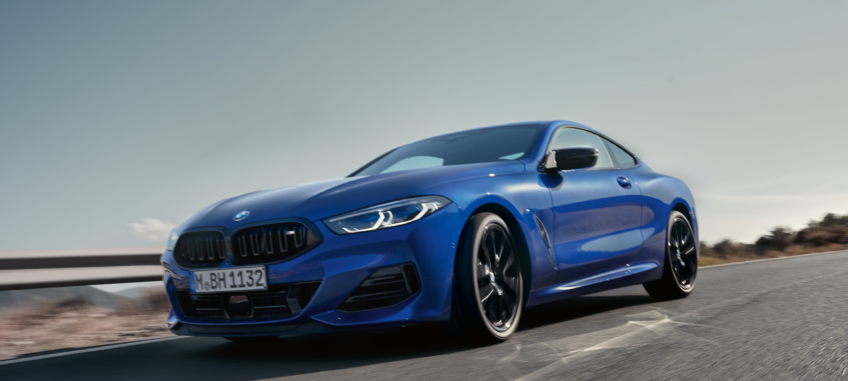 BMW 8-serie Coupé G15 LCI modellomarbetning 2022 BMW M850i xDrive M Portimao Blå metallic sedd framifrån ur grodperspektiv körande BMW 8-serie Coupé G15 LCI modellomarbetning 2022 BMW M850i xDrive M Portimao Blå metallic sedd framifrån ur grodperspektiv körande på landsväg med kuperat landskap i bakgrunden