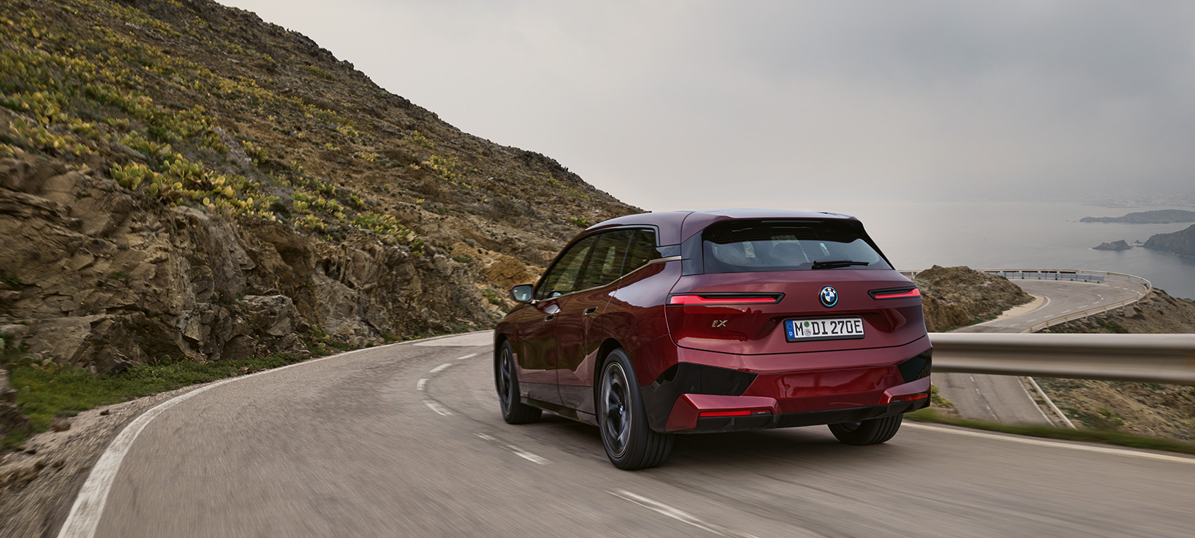 BMW iX i20 2021 eldriven SUV BMW iX xDrive50 körandes på en kustväg BMW iX i20 2021 eldriven SUV BMW iX xDrive50 aventurinröd sedd bakifrån körandes på en kustväg