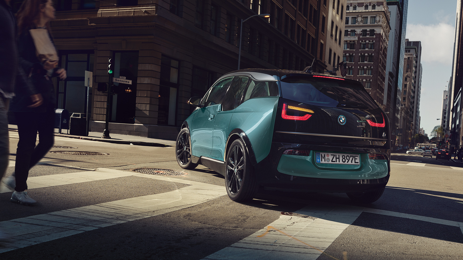 BMW i3 2018 sedd snett bakifrån körandes BMW i3 I01 2018 Jucarobeige med accent Frozen Grey metallic sedd snett bakifrån körandes i stadskuliss