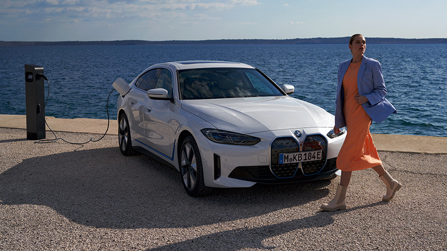 BMW i4 eDrive40 G26 2021 sedd snett framifrån vid laddningsstation Kvinna framför BMW i4 eDrive40 G26 2021 Mineralvit sedd snett framifrån intill laddstolpe vid havet