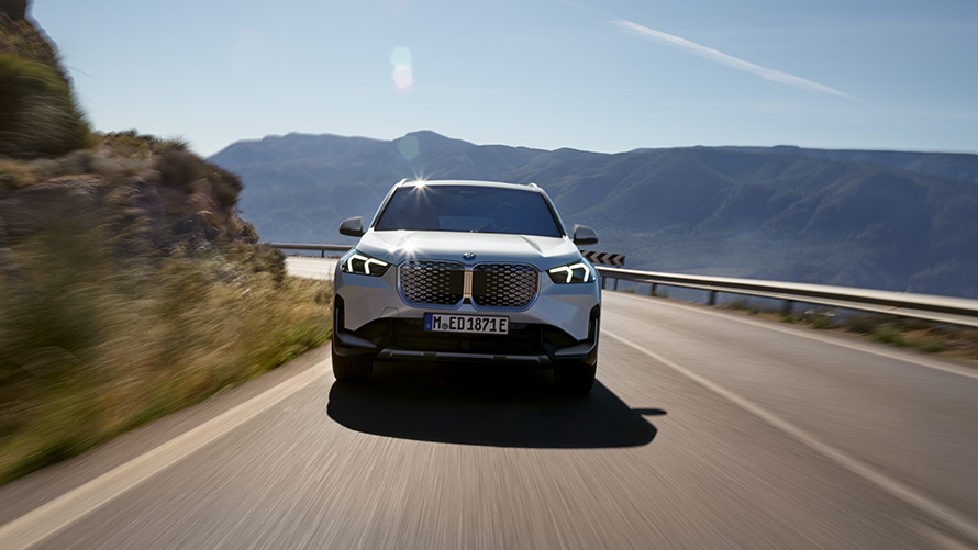 BMW iX1 U11 BEV 2022 framifrån BMW iX1 U11 BEV 2022 Mineralvit metallic sedd körandes framifrån på en bergsväg