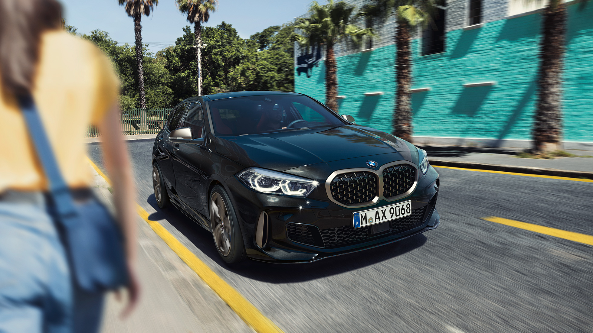 BMW M135i xDrive, trefjärdedelsvy framifrån. BMW M135i xDrive i safirsvart metallic, trefjärdedelsvy framifrån, körande med kvinna i förgrunden.