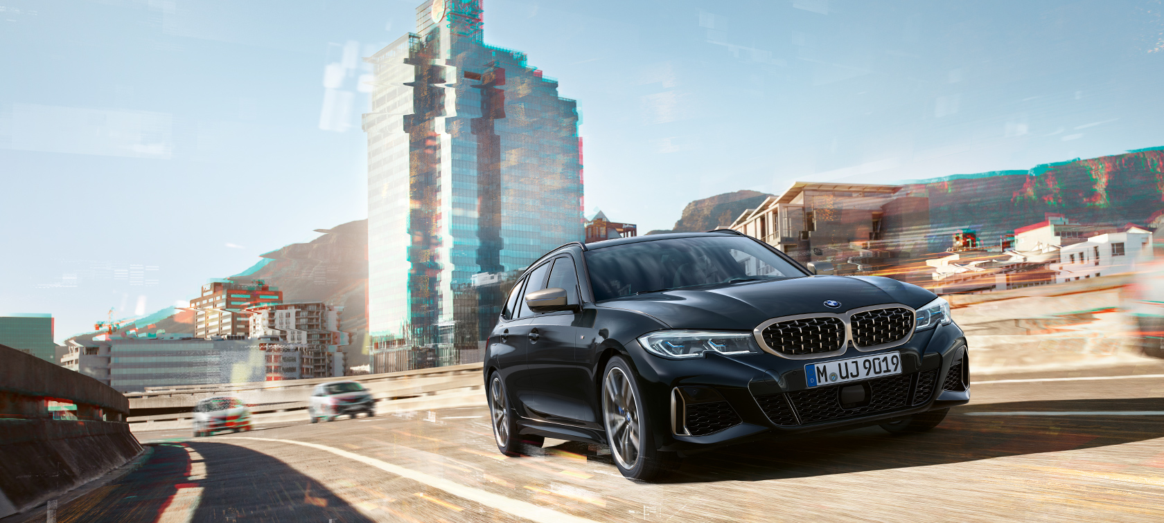 Trefjärdedelsvy framifrån körandes framför stadskuliss BMW M340i xDrive Touring, trefjärdedelsvy framifrån, körandes framför stadskuliss