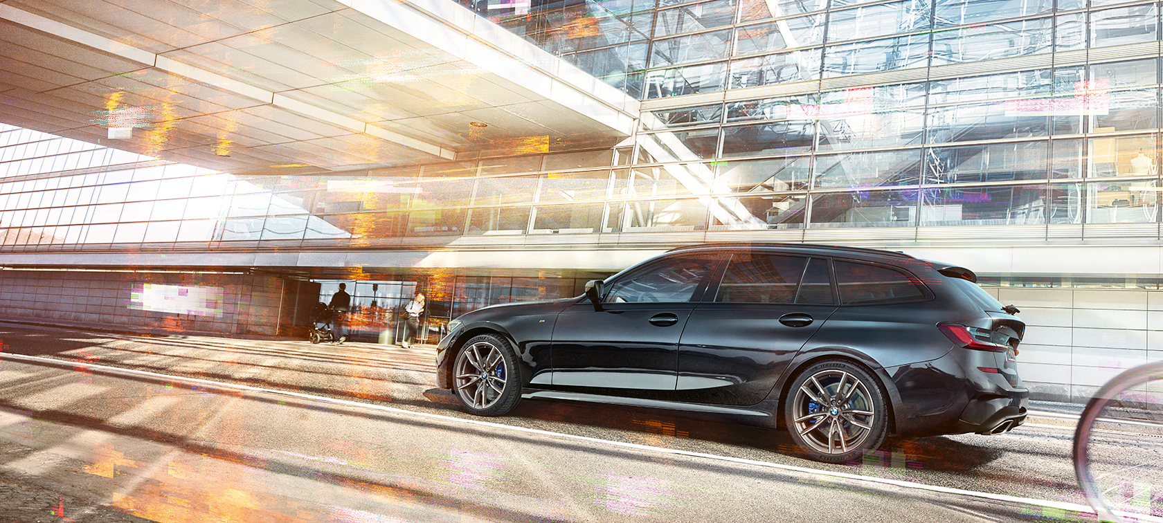 BMW M340i xDrive Touring, sedd från sidan körandes BMW M340i xDrive Touring, sedd från sidan körandes framför glasbyggnad i stadsmiljö