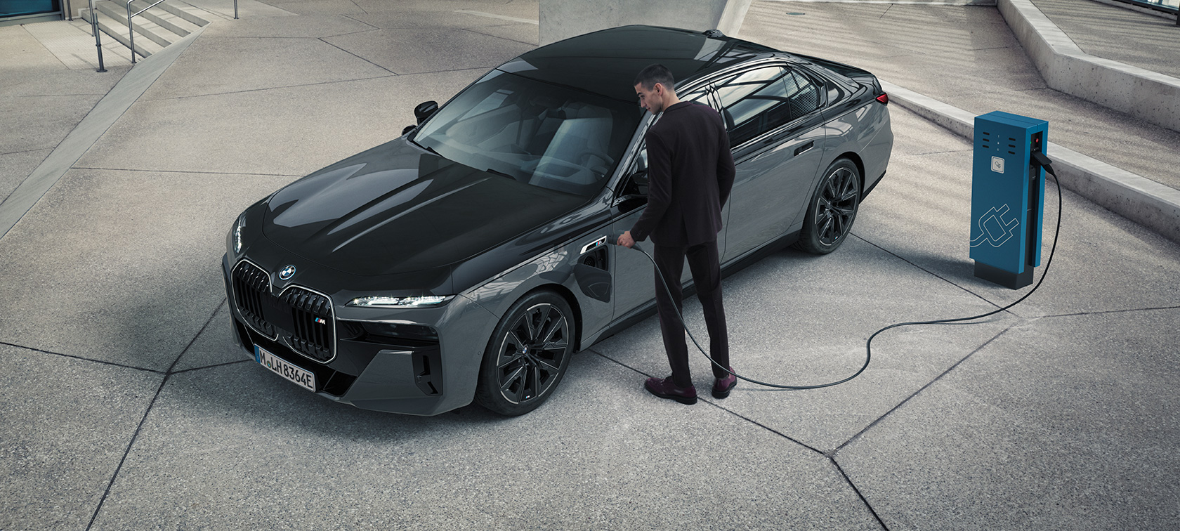 BMW M760e xDrive Sedan G70 M Performance-paket BMW Individual tvåtonslack Safirsvart metallic BMW Individual Dravitgrå metallic sedd snett framifrån man med laddningskabel intill laddstation