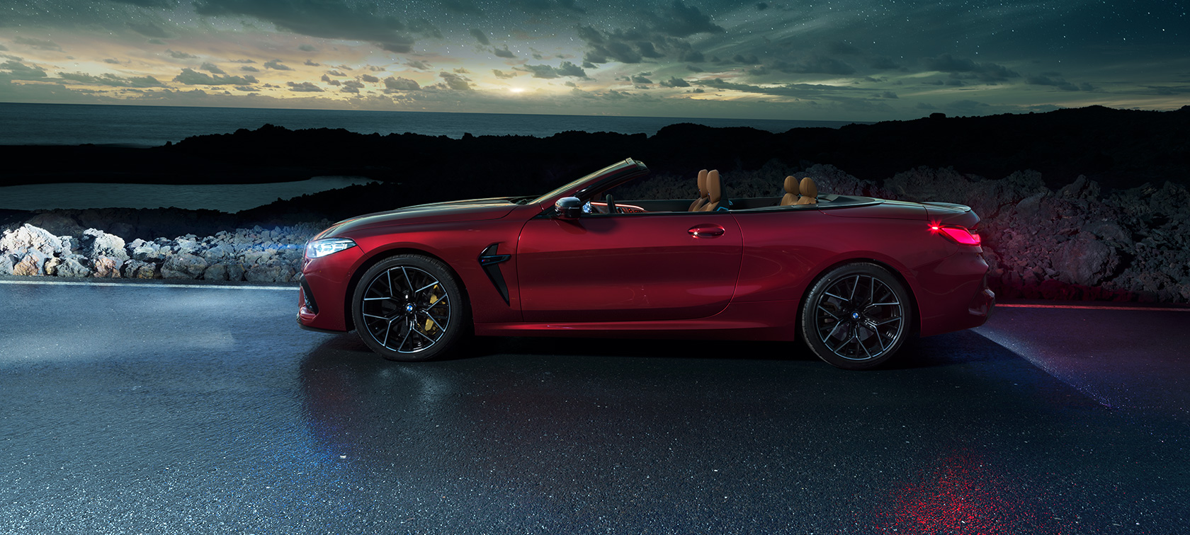 BMW M8 Competition Cabriolet, sedd från sidan. BMW M8 Competition Cabriolet i Motegi Rödmetallic med öppen sufflett, sedd från sidan, parkerad under stjärnhimmel vid kustväg.