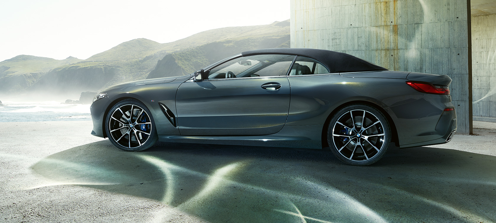 BMW M850i xDrive Cabriolet, sedd från sidan. BMW M850i xDrive Cabriolet i Dravit Gråmetallic med stängd sufflett, sedd från sidan framför en ås.