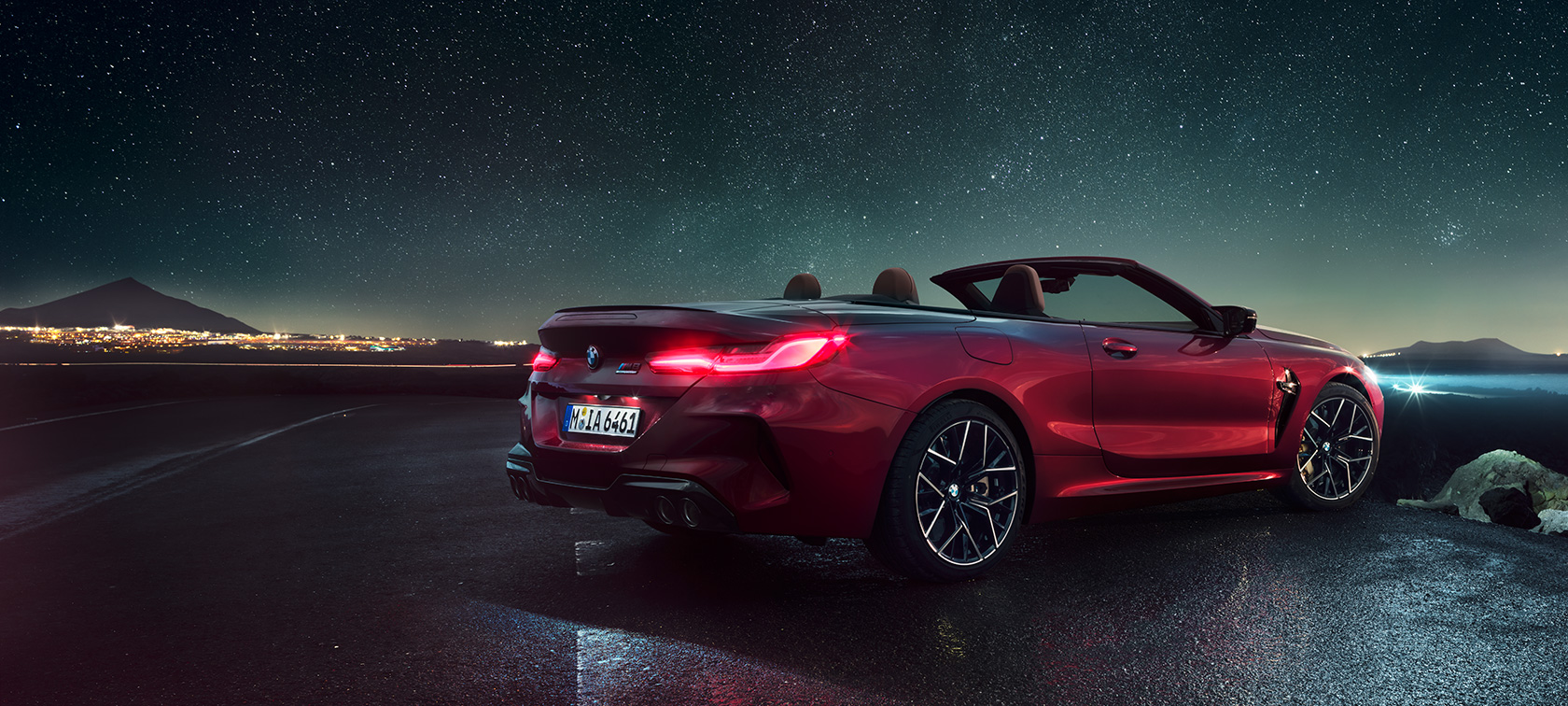 BMW M8 Competition Cabriolet design trefjärdedelsvy bakifrån. BMW M8 Competition Cabriolet i Motegi Rödmetallic med öppen sufflett, trefjärdedelsvy bakifrån, parkerad med man under stjärnhimmel.