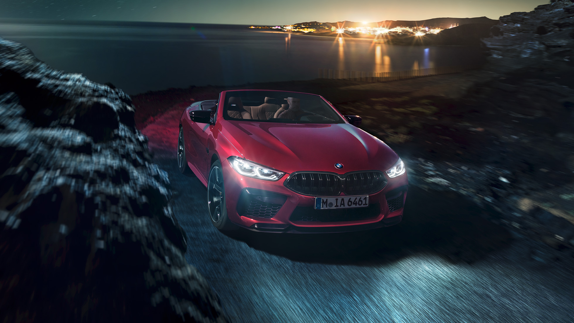BMW M8 Competition Cabriolet design trefjärdedelsvy framifrån. BMW M8 Competition Cabriolet i Motegi Rödmetallic med öppen sufflett, trefjärdedelsvy framifrån, körandes nattetid på en väg vid kusten.