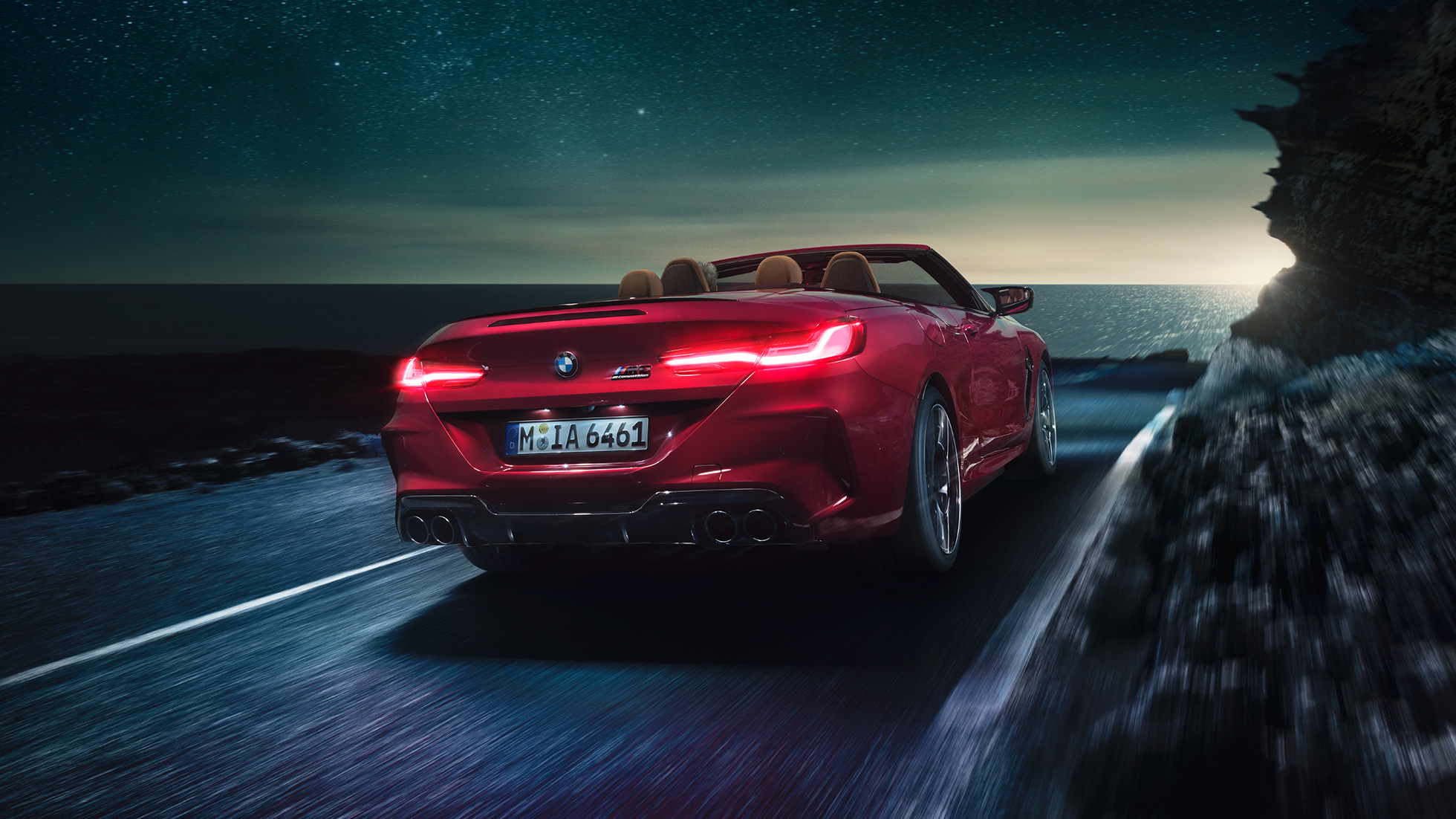 BMW M8 Competition Cabriolet design trefjärdedelsvy bakifrån. BMW M8 Competition Cabriolet i Motegi Rödmetallic med öppen sufflett, trefjärdedelsvy bakifrån, körandes nattetid på en väg vid kusten.