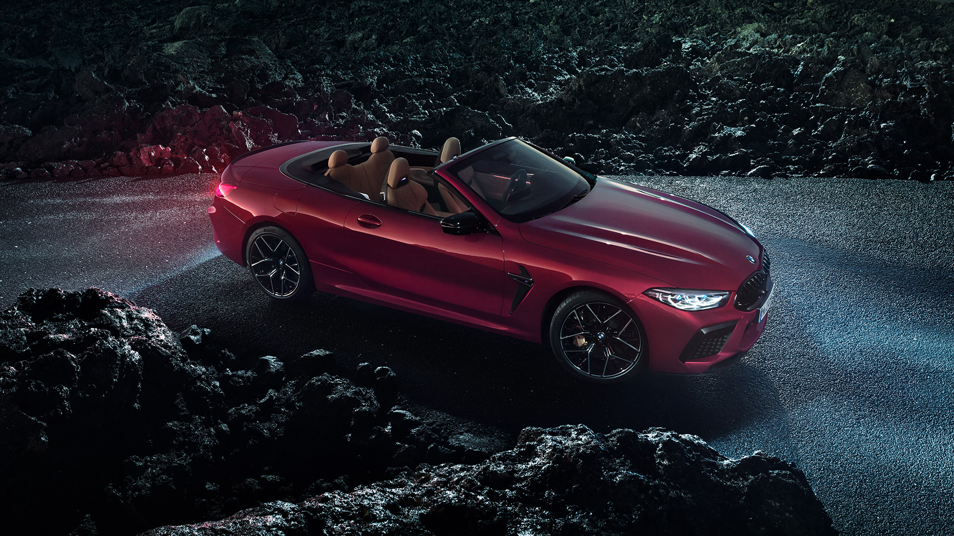BMW M8 Competition Cabriolet design trefjärdedelsvy från sidan. BMW M8 Competition Cabriolet i Motegi Rödmetallic med öppen sufflett, trefjärdedelsvy från sidan, parkerad nattetid på en asfalterad väg.