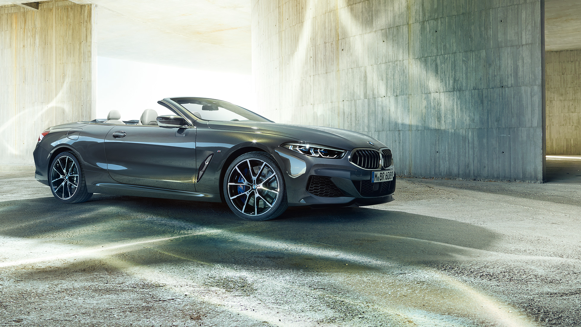 BMW M850i xDrive Cabriolet, trefjärdedelsvy från sidan. BMW M850i xDrive Cabriolet i Dravitgrå metallic med öppen sufflett, trefjärdedelsvy från sidan, parkerad framför betongkomplex.