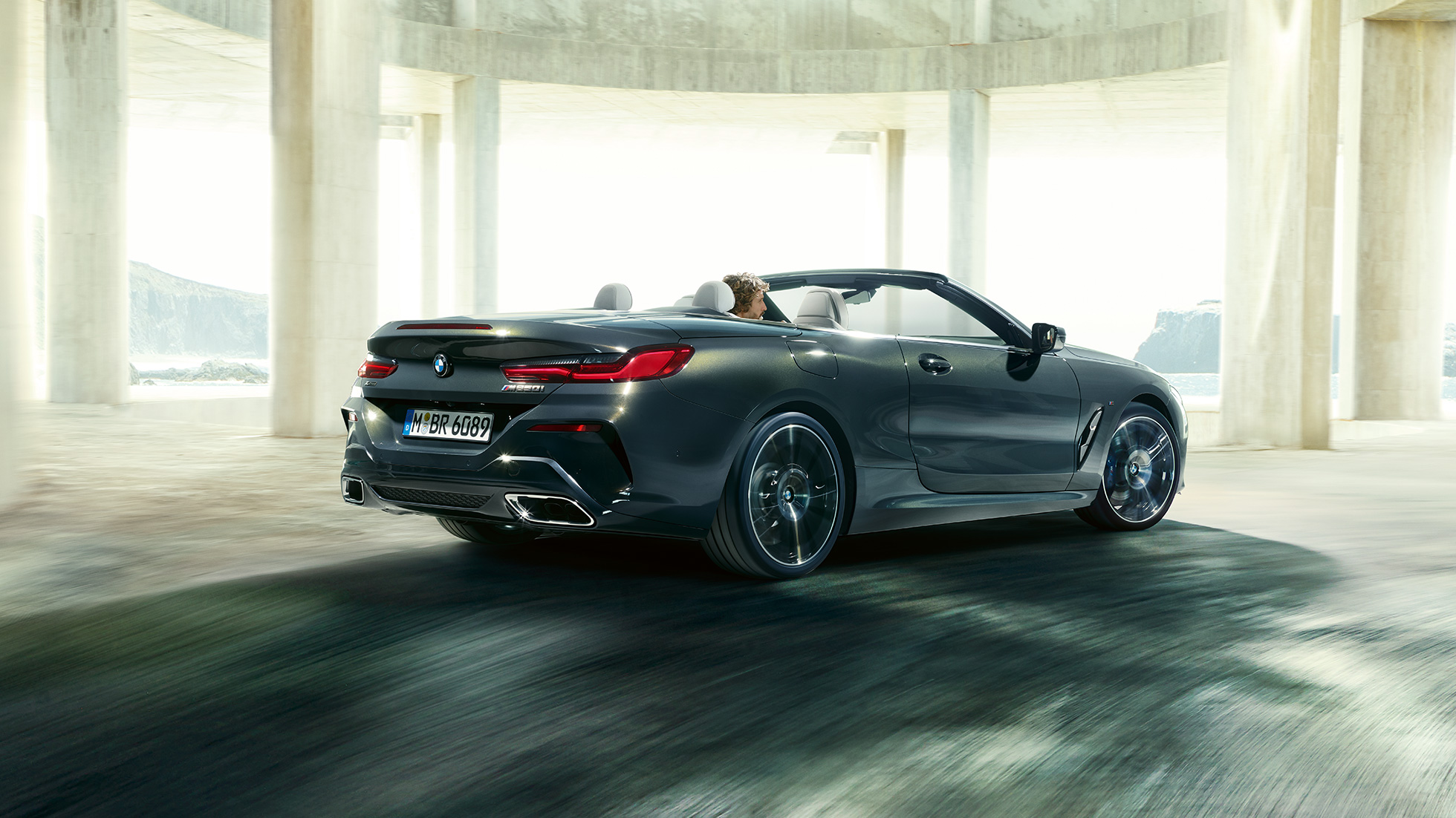 BMW M850i xDrive Cabriolet, trefjärdedelsvy bakifrån. BMW M850i xDrive Cabriolet i Dravitgrå metallic med öppen sufflett, trefjärdedelsvy bakifrån, körandes framför betongkomplex.