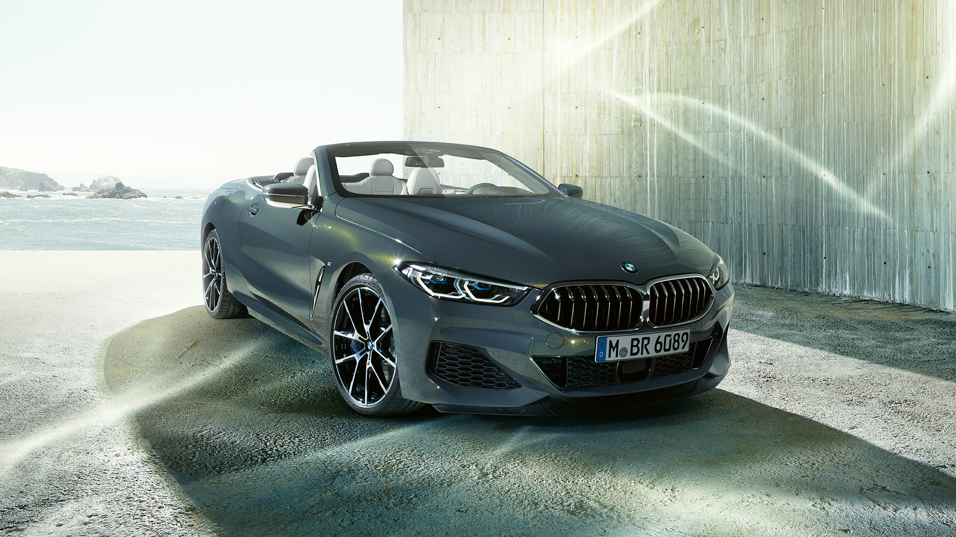 BMW M850i xDrive Cabriolet, trefjärdedelsvy framifrån. BMW M850i xDrive Cabriolet i Dravitgrå metallic med öppen sufflett, trefjärdedelsvy framifrån, parkerad framför kuststräcka.