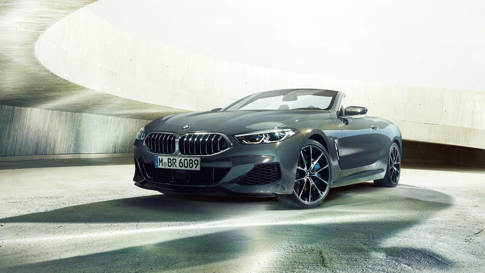 BMW M850i xDrive Cabriolet, trefjärdedelsvy framifrån. BMW M850i xDrive Cabriolet i Dravitgrå metallic med öppen sufflett, trefjärdedelsvy framifrån, parkerad framför betongkomplex.