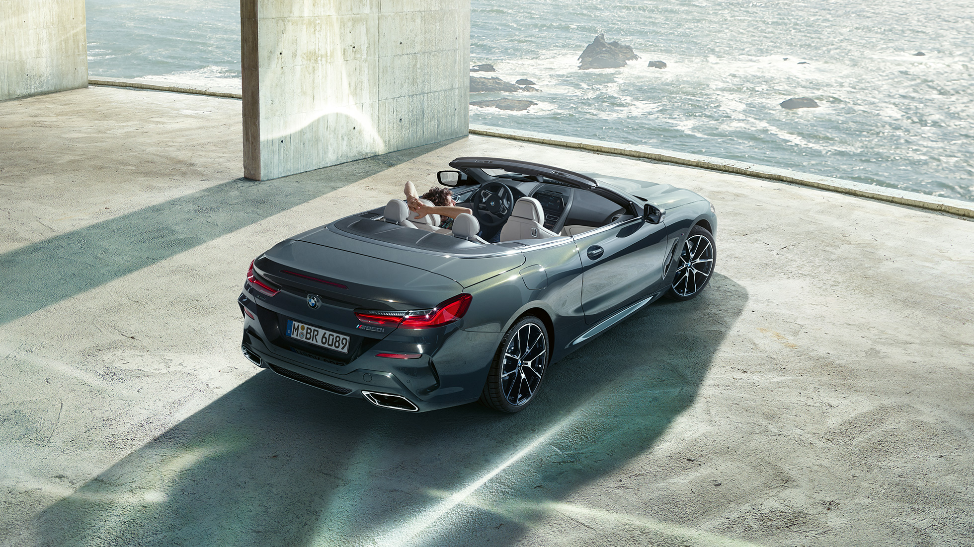 BMW M850i xDrive Cabriolet, fågelperspektiv. BMW M850i xDrive Cabriolet i Dravitgrå metallic med öppen sufflett, sedd ur fågelperspektiv, parkerad framför betongkomplex vid havet.