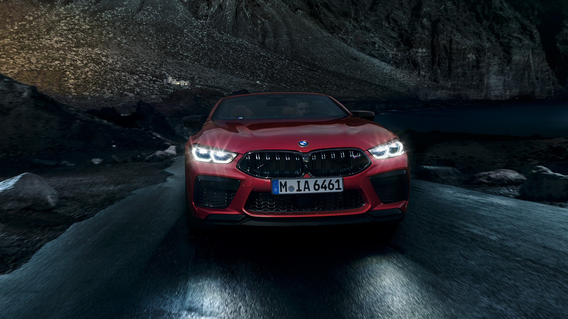 BMW M8 Competition Cabriolet, sedd framifrån. BMW M8 Competition Cabriolet i Motegi Rödmetallic med tända strålkastare, sedd framifrån, körandes nattetid.