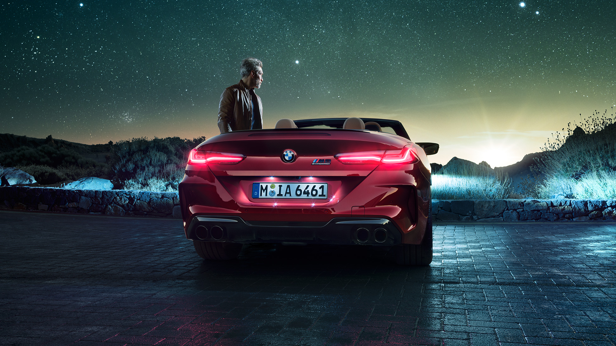 BMW M8 Competition Cabriolet, sedd bakifrån. BMW M8 Competition Cabriolet i Motegi Rödmetallic med öppen sufflett, trefjärdedelsvy bakifrån, parkerad med man under stjärnhimmel.