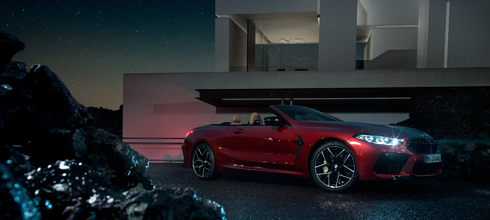 BMW M8 Competition Cabriolet design trefjärdedelsvy från sidan. BMW M8 Competition Cabriolet i Motegi Rödmetallic, trefjärdedelsvy från sidan, parkerad framför villa på natten.