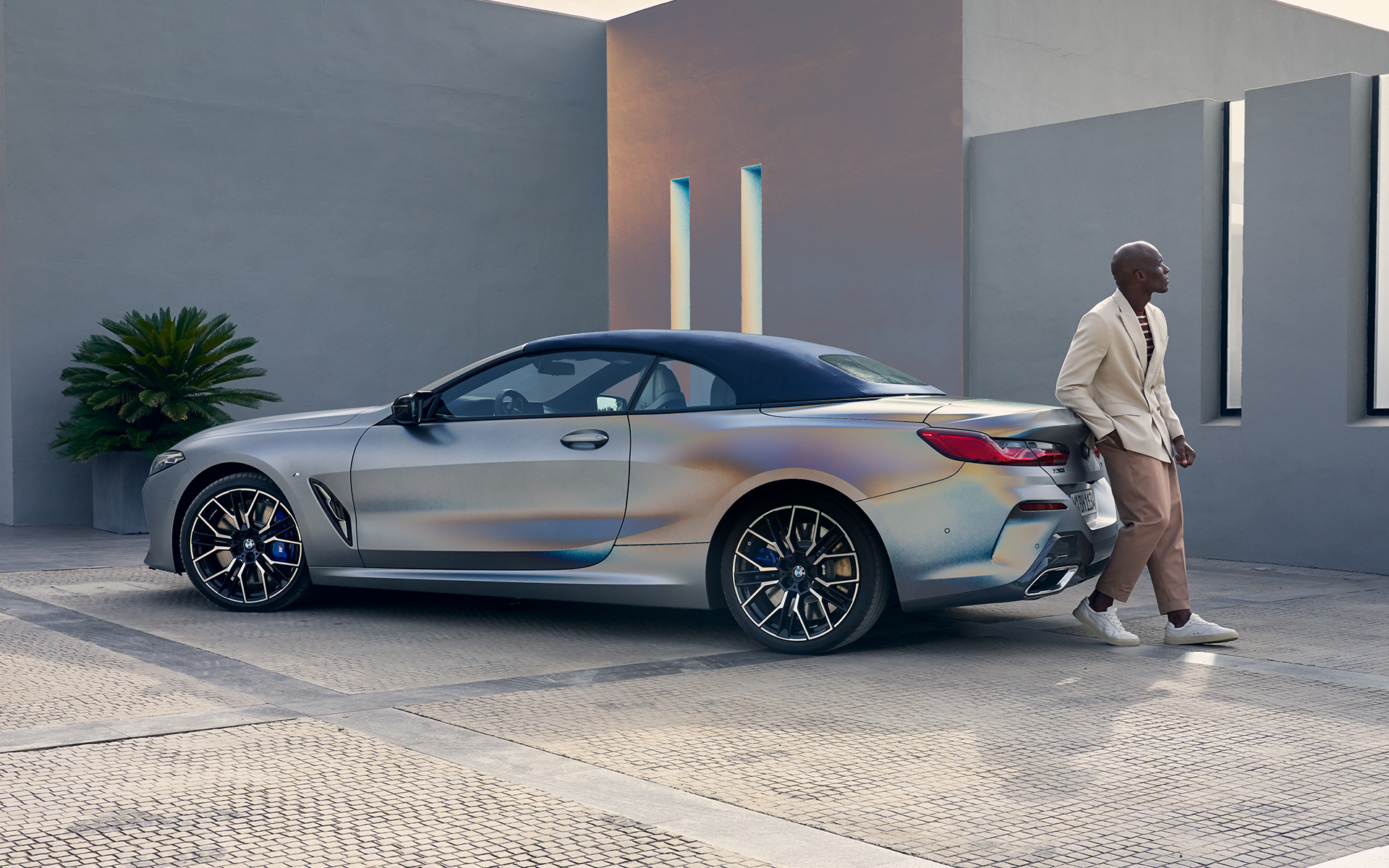 Snett bakifrån BMW M850i xDrive Cabriolet G14 LCI Facelift 2022 BMW M850i xDrive Cabriolet G14 LCI Facelift 2022 BMW Individual Frozen Pure Grey metallic sedd snett bakifrån med manlig modell som lutar sig mot bilen