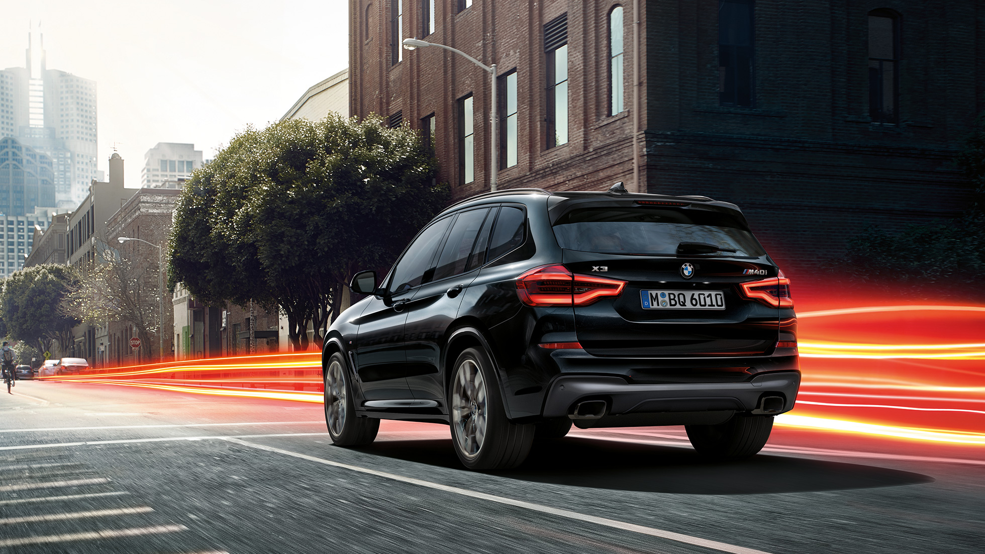 BMW X3 M40i, Trefjärdedels vy bakifrån. BMW X3 M40i i Safirsvart metallic, kommer körande, Exteriör, trefjärdedels vy bakifrån framför stadskuliss med ljusstrimmor.