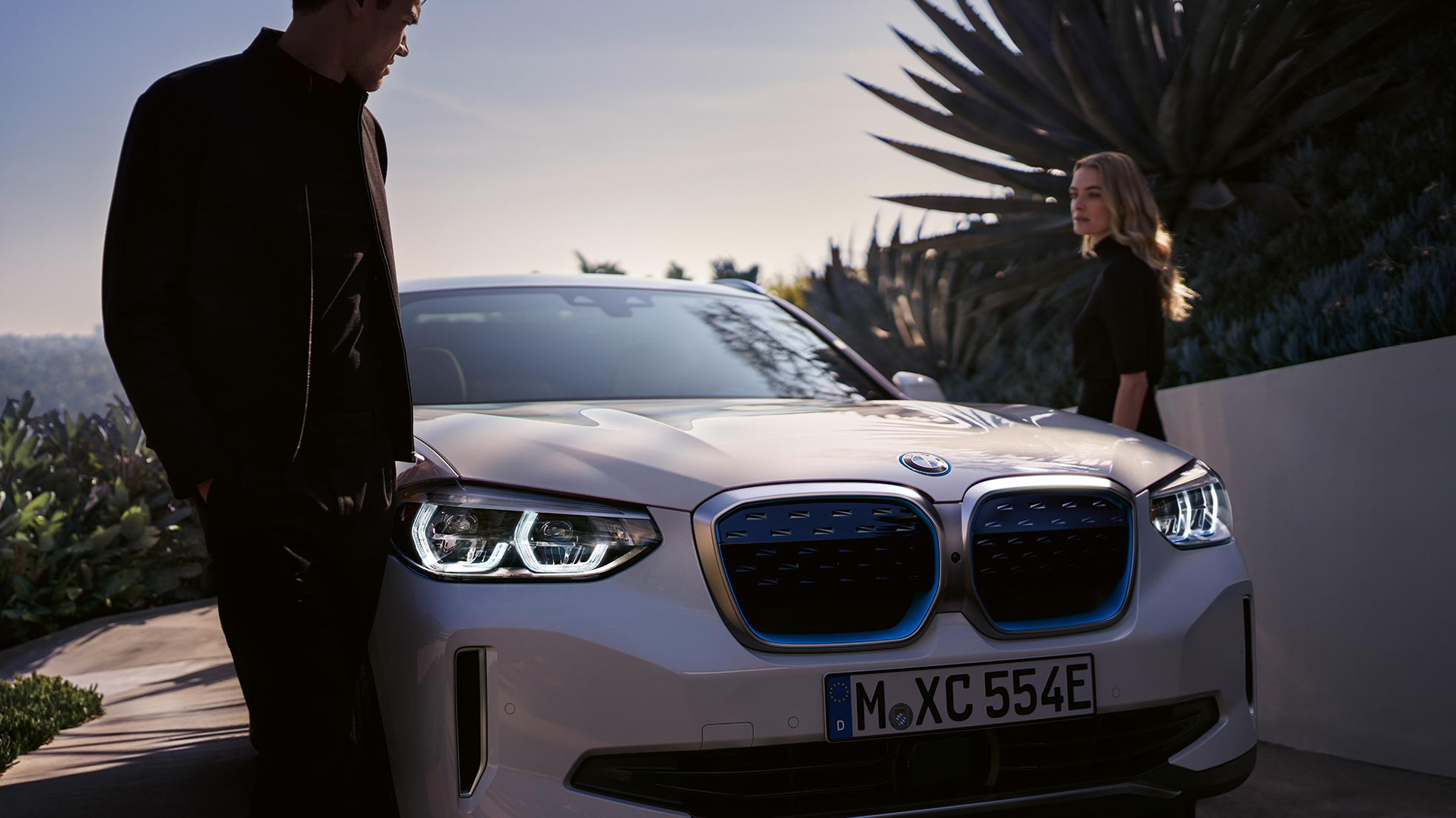 BMW iX3 G08 sedd snett framifrån med BMW i njurar BMW iX3 G08 2020 Mineralvit metallic sedd snett bakifrån med kvinna med BMW i njurar