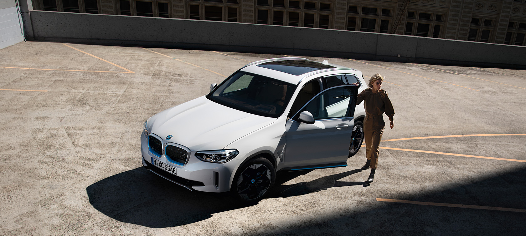 BMW iX3 G08 sedd snett framifrån kvinnlig förare BMW iX3 G08 2020 Mineralvit metallic sedd snett bakifrån med kvinna med stad i bakgrunden