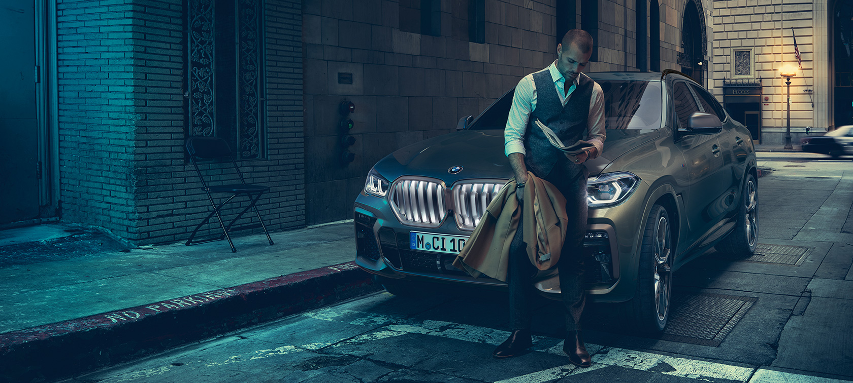 En man lutar sig mot en BMW X6: en man står lutad mot en BMW X6, sedd från sidan framför stadskuliss.
