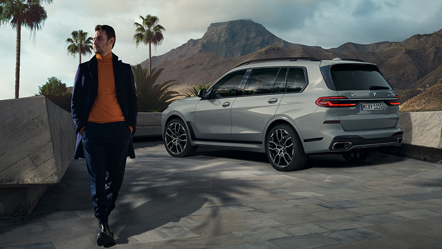 Man som står framför parkerad BMW X7 (G07) Remote Control Parking Remote Control Parking man som står framför parkerad BMW X7 (G07) sedd snett bakifrån bland palmer med bergslandskap i bakgrunden