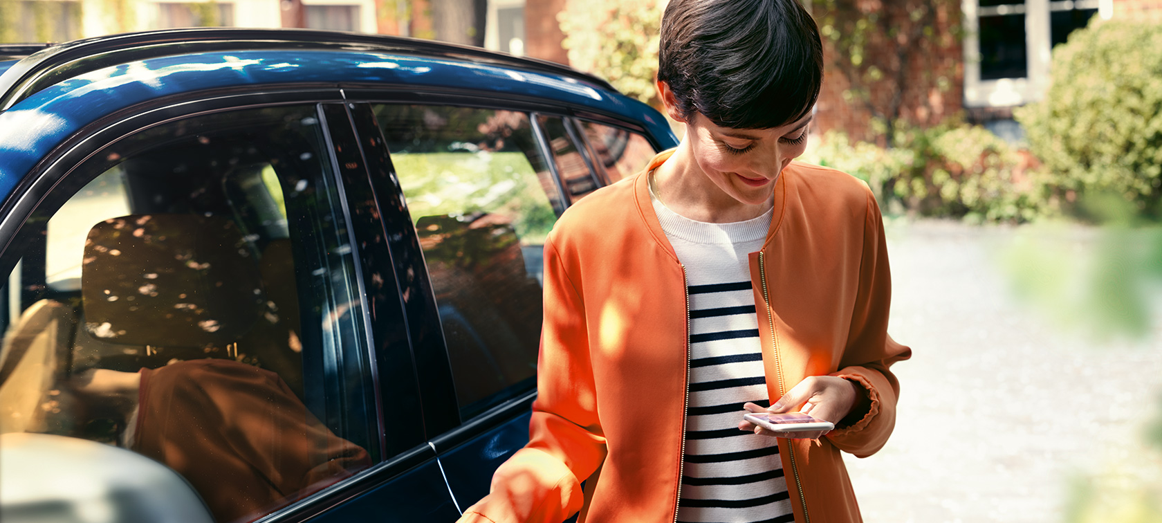 En mörkhårig kvinna framför en BMW En mörkhårig kvinna med orange jacka står framför en BMW och tittar på sin telefon