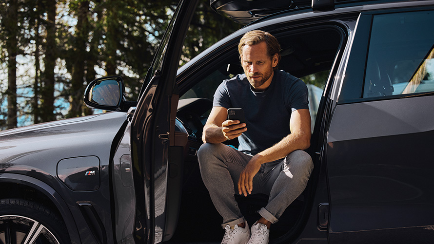 En man sitter i en BMW och tittar i sin smartphone BMW Vår- och sommartillbehör - en man sitter i en BMW och tittar i sin smartphone