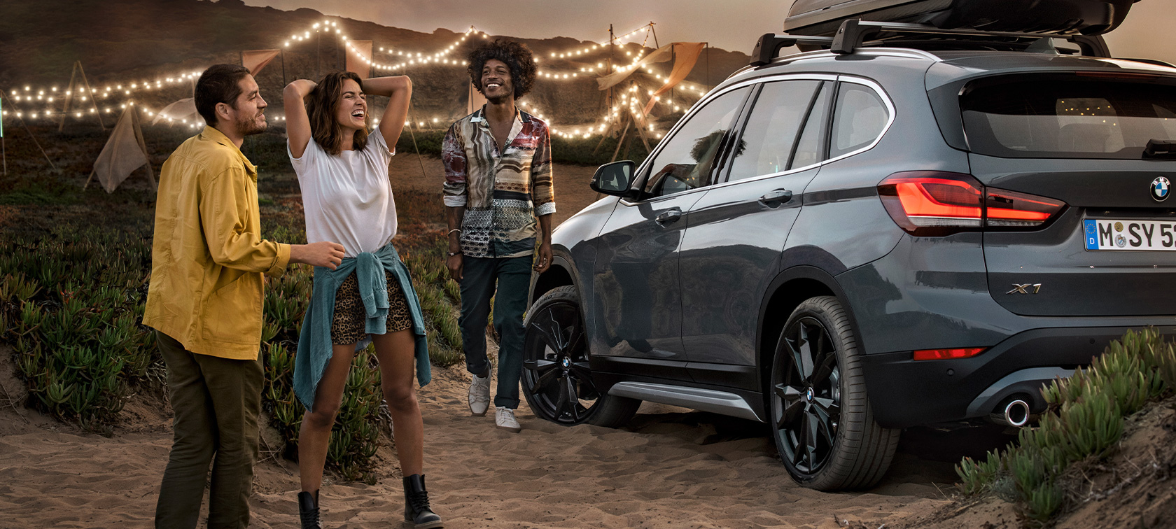 Jublande trio i BMW X1 med BMW erbjudande sommar Lycklig trio i mineralgrå BMW X1 utrustad med BMW erbjudande sommar trefjärdedelsvy bakifrån på stranden